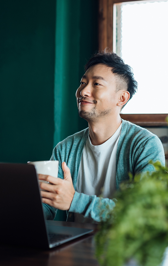Person sitting at a table with a laptop, holding a white mug and looking slightly upward. Background features a green wall and a window with natural light, with greenery visible in the foreground.